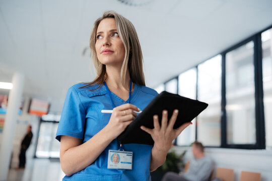 Portrait of confident female doctor standing in Hospital corridor. Beautiful nurse wearing blue scrubs, holding clipboard standing in modern private clinic, low angle shot.