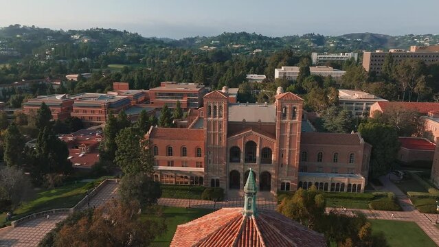 Aerial view of UCLA campus with Royce Hall center stage, Romanesque architecture, green spaces, and Westwood's urban backdrop in golden morning - afternoon light.