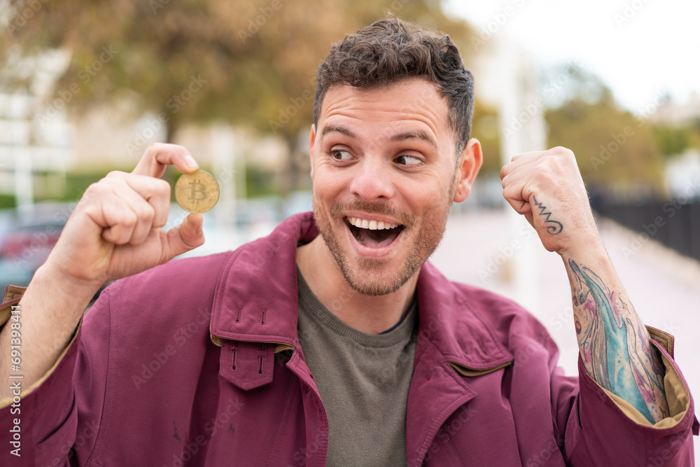 Wall mural young caucasian man holding a bitcoin at outdoors celebrating a victory