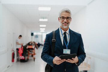 Portrait of pharmaceutical sales representative in medical building, waiting for doctor, presenting new pharmaceutical product. Smiling drug rep standing in hall holding tablet. - obrazy, fototapety, plakaty
