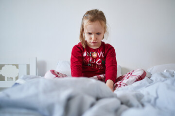 Unhappy preschooler girl wearing Christmas pajamas, sitting on bed