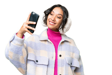 Young Argentinian woman wearing winter muffs over isolated background making a selfie