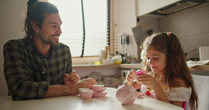 Happy brunette man in green checkered shirt. Lonely father plays tea party game with his little daughter brunette girl in pink dress using toy pink cups and kettle in kitchen