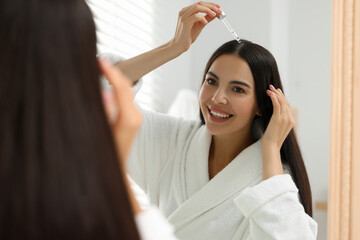 Beautiful woman applying hair serum indoors, reflection in mirror. Cosmetic product