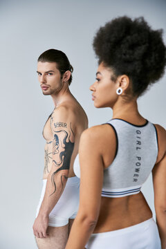 focus on tattooed man with ponytail posing next to his blurred girlfriend and looking at camera