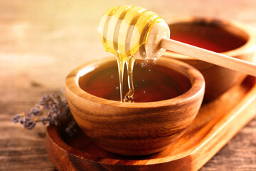 Natural honey dripping from dipper into wooden bowl on table under sunlight, closeup