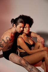 Fototapeta premium loving multicultural couple sitting on floor alluringly together and hugging warmly on gray backdrop