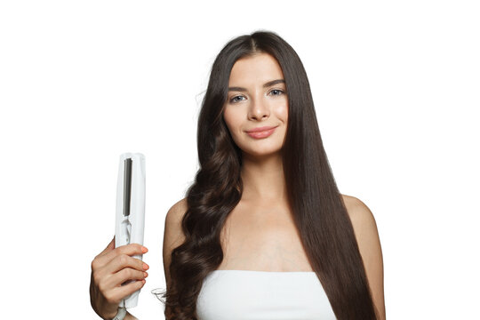 Cheerful smiling woman using hair iron and straightening her long brown hair on white background, studio fashion beauty portrait. Hair ironing and hairstyle concept