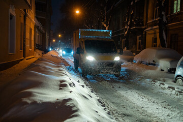 Yellow lorry van moving at night in the heavy snowfall on a snowy street with lights on - obrazy, fototapety, plakaty