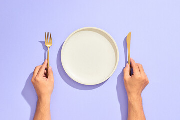 Dinner place setting. White empty plate and hand holding gold fork and knife on lilac background