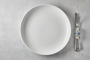 Empty white dinner plate and cutlery wrapped in a hundred dollar bill on a gray stone table. Top view with copy space