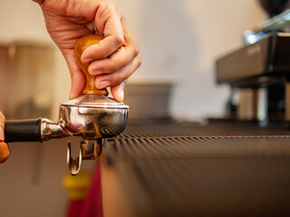 Hand of a barista holding a portafilter and a coffee tamper making an espresso coffee. Barista presses ground coffee using a tamper in a coffee shop