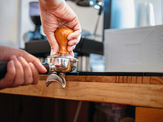Hand of a barista holding a portafilter and a coffee tamper making an espresso coffee. Barista presses ground coffee using a tamper in a coffee shop