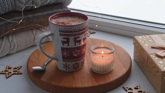 Winter windowsill still life. Red ceramic cup of hot coffee on window sill. Christmas decorations on the background. Cozy home picture. Warm woolen knitted sweaters, Burn Candle, Cookies. Stock photo	