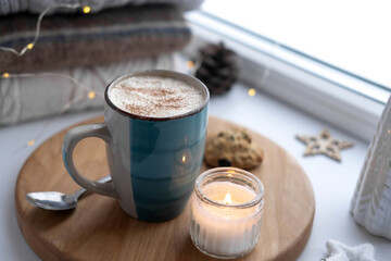 Winter windowsill still life. Blue ceramic cup of hot coffee on window sill. Christmas decorations on the background. Cozy home picture. Warm woolen knitted sweaters, Burn Candle, Cookies. Stock photo