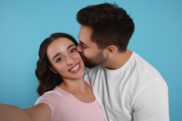 Man kissing his smiling girlfriend while she taking selfie on light blue background