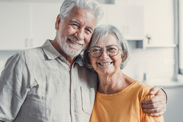 Headshot portrait of smiling elderly 60s husband and wife sit relax on couch hugging cuddling, happy mature old couple rest on sofa in living room embrace look at camera show love and care.