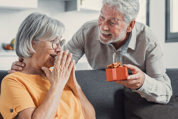 Smiling senior husband making surprise giving gift box to amazed wife, happy aged woman get...