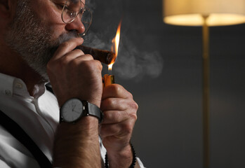 Bearded man lighting cigar indoors, closeup. Space for text