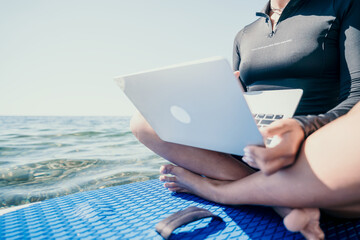 Woman sea laptop. Digital nomad, freelancer with laptop working on sup board at calm sea beach....