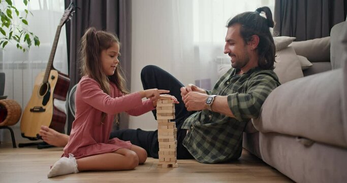 A lonely father, a brunette man in a Green checkered shirt plays with his little daughter, a brunette girl in a pink dress, in the board game Jenga, leaning on a gray sofa on the floor in a modern