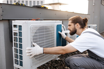 Adult bearded construction worker crouching and keeping hands on air conditioner while checking...