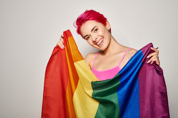 portrait of smiling and young woman with pink hair posing with lgbt rainbow flag on grey backdrop
