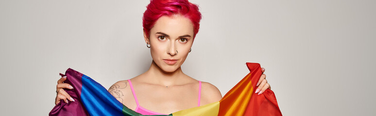 Naklejka premium portrait of tattooed young woman with pink hair posing with lgbt rainbow flag on grey, banner