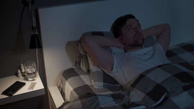 Young man looks at the ceiling at night while lying in bed instead of sleeping, checking the time on his mobile phone, suffering from insomnia. Displeased male lying in bed unable to fall asleep.