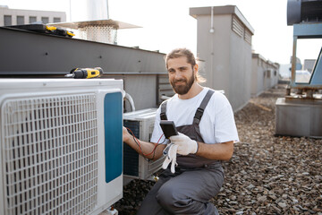 Concentrated man squatting down and holding voltage tester while applying to air conditioner. Builder man closed in overalls performing work and checking serviceability of cooling system.
