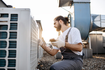 Side view of professional builder sitting in front of air conditioner and using measuring device on roof of factory. Caucasian focused male with braided hair testing voltage of climate control unit.