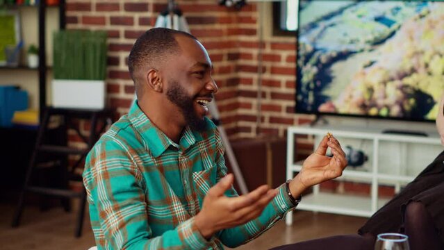 Smiling African American Man Having Interesting Conversation With BIPOC Apartment Party Guests While Eating Snacks. Close Up On Charcuterie Board On Table Next To Inclusive Group Of Friends Talking