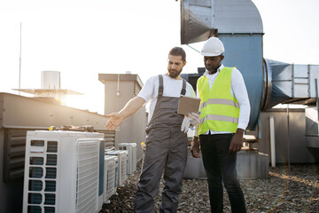 African american supervisor and caucasian technician standing next to each other and looking at modern tablet screen. Qualified factory workers reading information about air conditioners on rooftop.