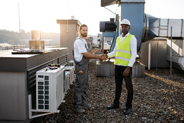 Serious engineers standing and shaking hands while working together on roof. Multicultural men dressed in construction uniforms completing matter with repair of appliances on fresh air. - obrazy, fototapety, plakaty