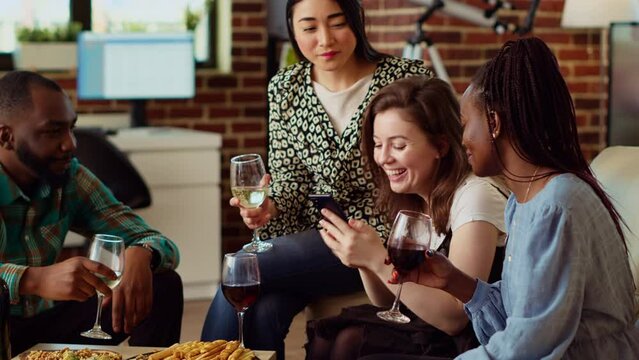 Diverse friends at apartment party using smartphone to laugh at social media posts made by acquaintance. Amused guests in living room gossiping about cringe images posted online by mate