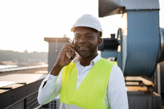 Portrait Of Male Worker Dressed In Work Uniform Standing And Smiling While Talking On Digital Mobile On Fresh Air. African American Man Having Conversation And Working With Devices On Roof Of Factory.