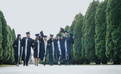 University students celebrating graduation in a park. Wearing gowns and caps, they congratulate...