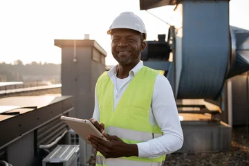 Foto op Canvas Serious factory worker standing and holding modern tablet in hands while servicing devices at factory. African american man wearing uniform inspecting area while performing work in local roof. © sofiko14