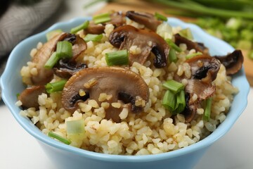 Delicious bulgur with mushrooms and green onion in bowl on table, closeup
