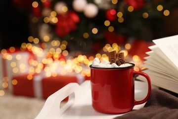 Delicious cocoa with marshmallows, cinnamon sticks and anise star in cup on white tray against blurred Christmas lights, closeup. Space for text