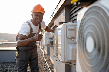 African american factory worker in orange hard hat standing and holding hands on air conditioner...