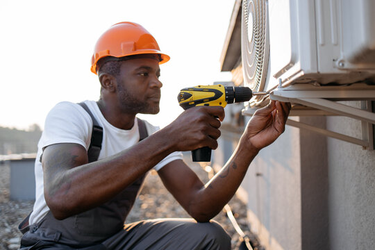 Portrait of handsome african american male using electrical screwdriver for repairing ventilation system outdoors. Smiling man with protective helmet looking directly and professionally using tool.