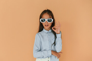 Okay hand gesture, in sunglasses. Cute young girl is in the studio against background