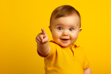 Curious baby opens its mouth in astonishment, standing on a solid bright yellow background, pointing a chubby finger to the left - obrazy, fototapety, plakaty