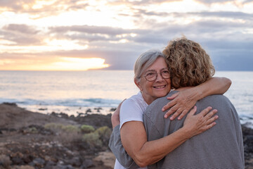 Fototapeta premium Romantic senior couple of women embrace each other on the seashore at sunset light, two embraced people stay together expressing love and tenderness