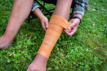 A person s leg being bandaged with an orange bandage. The person is sitting on a green folding chair on a grassy lawn. The bandage is being applied to the lower leg and ankle area.