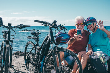 Video call concept. Happy senior cyclist couple at the beach with electric while using mobile phone...