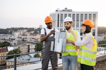 Friendly team of three project managers inspecting reduced copy of building that holding one of workers on rooftop with beautiful landscape on background. Concept of architecture and construction.