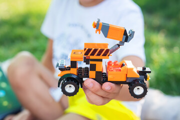 A child s hands holding a toy truck made of building blocks. The truck is orange and black with stripes on the side. The child is sitting on a grassy lawn in a park on a sunny day.