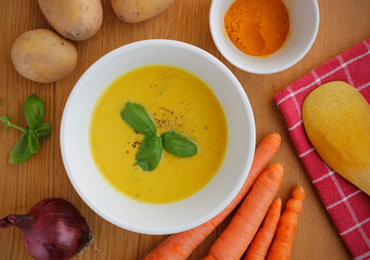 White bowl of potato and carrot cream soup with ingredients, wooden spoon and red kitchen towel on wooden table. Healthy vegetarian soup. Top view food. 